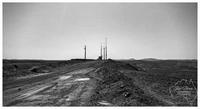 A low angle, black and white photograph signed by Kev Peirce. A rough, dirt track littered with rubble leads up a steep embankment toward a cluster of utility poles and a radio communication tower in the distance.  The foreground is heavily textured with shadows and loose rock. The horizon is wide and flat, with low hills visible in the far background under a bright, clear sky.  The landscape suggests an arid, remote, or mining affected area, likely near Leigh Creek.