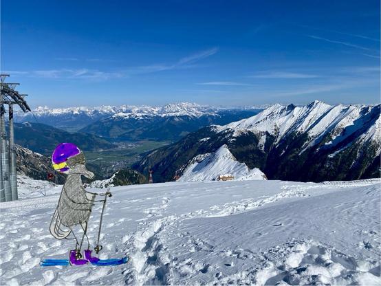 A cartoonish character resembling a fly with Ski goggles and a helmet stands on skis in real life photograph of a snowy mountain landscape. The background features towering snow-capped mountains under a clear blue sky. Ski lifts are visible on the left, and a small building is seen in the distance