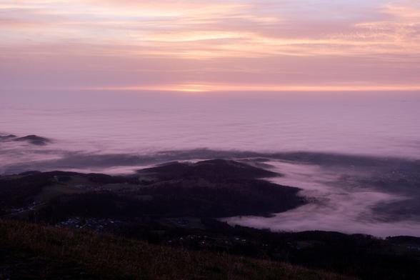 Blick von einem Berg ins Tal. Fast das gesamte Tal liegt unter einer dicken Nochnebeldecke. Nur im Vordergrund sieht man den Abhang und einige Hügel, die sich über die Nebeldecke herausschieben. An den Rändern gibt es leichte Nebelschwaben, die nach hinten hin dichter werden.