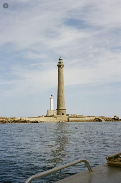 Leuchtturm auf einer kleinen Felsinsel vor blauem Himmel, mit einem zweiten, kleineren weißen Leuchtturm daneben; ruhiges Meer im Vordergrund, Blick von einem Boot mit Reling.
