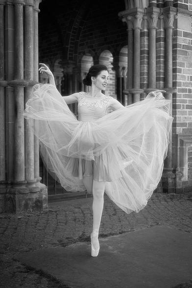 Eine Ballerina steht auf Zehenspitzen vor einer Backsteinmauer mit Säulen und Rundbögen. Sie trägt ein langes, fließendes Tüllkleid mit Spitzenapplikationen am Oberteil und hat die Arme anmutig erhoben, während der Rock ihres Kleides weit ausgebreitet ist.