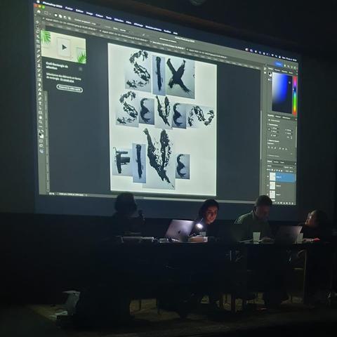 Photograph of a conference room in the dark and three people seen through their silhouettes on a long table, there's a projection on the wall where you can see a Photoshop window open and the text "Six Seven Five" spelled with some hand drawn experimental letters.