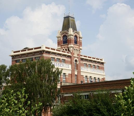 Bâtiment industriel du début du XXe siècle en brique rouge et pierre portant une enseigne « Brasseries Motte-Cordonnier », émergeant de la végétation. Une tourelle s’élève au milieu, ornée de vitraux portant une étoile à cinq branches. Des motifs blancs sur rouge viennent égayer la façade à plusieurs endroits.
