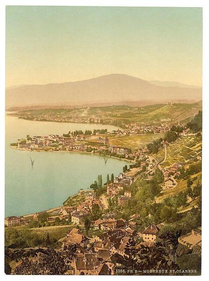 The image depicts a picturesque, aerial view over the town of Montreux and Clarens in Switzerland. It captures rolling green hillsides with patches of vineyards that gently slope down towards Lake Geneva below. The lake is dotted with boats sailing near what appears to be an anchored sailboat or windmill structure.

Settlements are densely packed along the shoreline, featuring buildings with red-tiled roofs typical of European architecture from this era. Some larger structures suggest institutional use, perhaps schools and churches, as indicated by their size and distinctive steepled rooftops which could signify a place of worship such as a church or chapel.

The surrounding area is rich in vegetation and trees dot the landscape around residential buildings. The town's layout reveals winding roads that connect various neighborhoods amidst this scenic environment.

In the distance are faint outlines of mountains, adding to the image’s serene setting. Above it all hangs a hazy sky with soft colors ranging from pale blue near the horizon transitioning into shades of orange and yellow higher up in the atmosphere, likely suggesting early morning or late afternoon light conditions.

The overall impression is one of historical charm combined with natural beauty, evoking an idyllic snapshot that might have been captured during a quiet day when life was perhaps simpler. The image carries an aura of tranquility and timelessness, s [...]