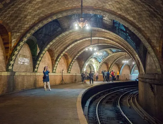 The image depicts an underground subway station with distinctive architectural features, including arches and brickwork. There are people present, some taking photos, while others appear to be gathered in groups. The lighting is warm, creating a cozy atmosphere typical of historic subway stations. The tracks are visible leading away from the platform area. Overall, it conveys a sense of place that is both historical and functional.

Image Credits: Wikimedia / Rhododendrites / CC BY-SA 4.0