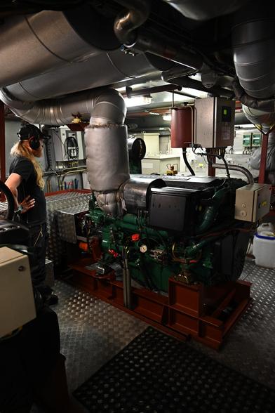 Blick in den Maschinenraum mit der Volvo Dieselmaschine von 2009, grün lackiert, mit zahlreichen Ventilen und Messinstrumenten. Der Auspuff ist dick isoliert, an der Decke des Raums Entlüftungsanlagen. Der Boden besteht aus Metall. Links der Maschinist mit Gehörschutz (wir durften für die Besichtigung auch nur mit Gehörschutz hinein). Der Raum war trotz der Isolierung noch gut warm.