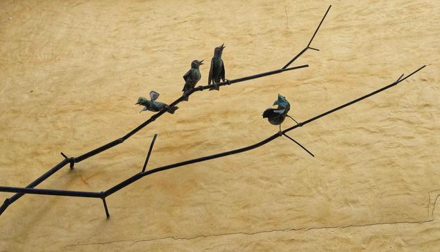 Four small, colourful metal sculptures of birds are perched on a minimalist, abstract black metal branch mounted against a textured yellow wall. The birds appear to be in various poses—some singing, some resting—adding a whimsical and artistic touch to the scene. The wall’s neutral, earthy tone contrasts with the dark, angular lines of the branch and the vibrant hues of the bird sculptures.