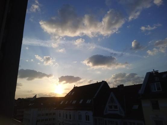 Foto im Querformat. Die Kamera blickt aus dem 5. Stock über Mietshäuser in der Düsseldorfer Helmholtzstraße. Der Blick geht nach Osten. Dort geht halb links gerade die Sonne auf, sie ist schon vollständig zu sehen. Der Himmel ist blau mit einigen teils auch dunkleren Wolken, um die Sonne herum ist er weiß-gelb.