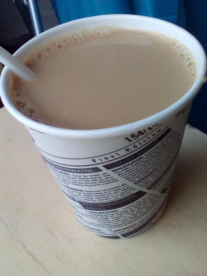 Foto im Hochformat. Die Kamera blickt von schräg oben auf einen Einweg-Kaffeebecher, der fast bis zum Rand mit Milchkaffee gefüllt ist. Der Becher steht auf einer hellen Holzoberfläche. Links ist noch ein kurzes Stück eines Plastiklöffels erkennbar, der aus dem Becher ragt.