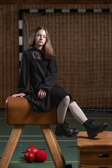 An 11-year-old girl is sitting on a brown leather-covered gym horse with wooden legs in a sports hall. She is wearing a black dress with lace detailing on the upper part, light-colored tights, and black lace-up boots. In the background, there is a handball goal with a net. Three red balls are lying on the gym floor in front of the gym horse.