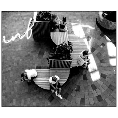 This black-and-white image shows an indoor space with a curved, circular brick-paved floor. In the center, there are two large, curved benches with wooden slats and green plants inside, viewed from above. Five people are present: two persons are sitting on the bench at the bottom of the image, two people are sitting on the top bench, another one on the right. Above the benches, a large, illuminated sign displays the word "info" in cursive writing. The area is well-lit with natural light streaming in from the top right. (draft by mistral.ai, revised by author)