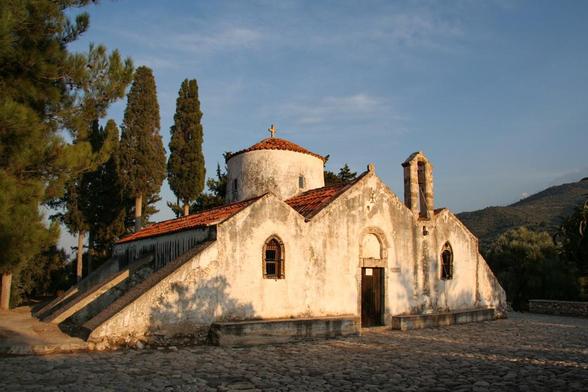 A small, weathered church with a distinctive dome and tiled roof, surrounded by tall cypress trees. The structure shows signs of age, with a rustic stone pathway leading to its entrance, set against a backdrop of hills under a clear sky.