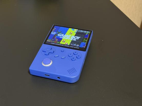 Sininen käsikokoinen pelikonsoli tummalla pöydän pinnalla; laitteessa kirkas näyttö, jossa lukee "GAME BOY ADVANCE", suuntapainike, analoginen tikku ja A/B/X/Y-painikkeet sekä USB- ja kuulokeliitännät.