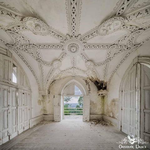 An ornate, white, abandoned room with detailed plaster ceiling, arched doors, peeling paint, and a partially collapsed wall, opening to a view of trees outside.