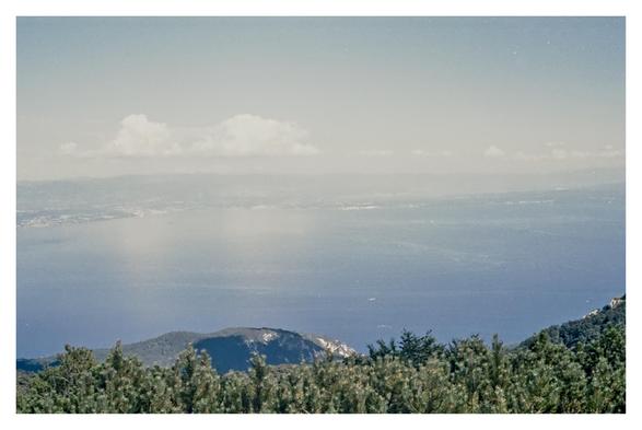 Color photograph of the Adriatic Sea, taken from Vojak, the highest peak in the Učka Mountains and in all of Istria at 1,401 meters above sea level. The image is rendered in light blue tones, with green treetops visible at the bottom, the Croatian coast in the background, the Adriatic Sea in between, and a pair of bright, shimmering clouds with tendrils stretching horizontally across the entire image above.

Translated with DeepL.com (free version)