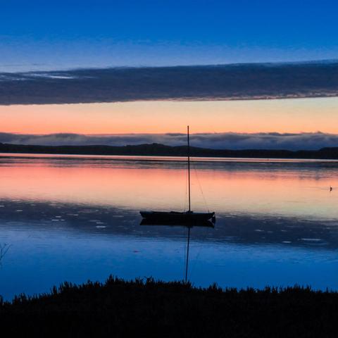 A wide, horizontal landscape photograph depicts a calm body of water at sunset. The sky is a gradient of orange and pink hues blending into a deep blue at the top of the frame. A dark silhouette of a small boat with a single mast is centered in the foreground, its reflection mirrored perfectly on the still water's surface. A dark, undefined landmass is visible along the distant horizon. The immediate foreground features a dark, blurred line representing vegetation, likely grass or reeds, along the water’s edge.