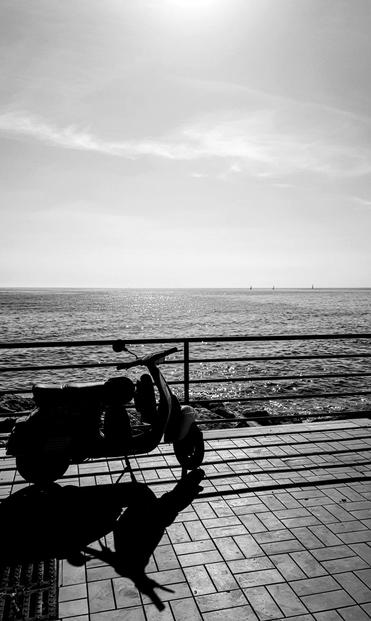 Foto verticale in bianco e nero scattata in Puglia: in primo piano una Vespa vista di lato, in silhouette, parcheggiata su una passeggiata piastrellata con ringhiera. Il sole alto crea un forte controluce e una lunga ombra dello scooter sul pavimento. Sullo sfondo il mare brilla.