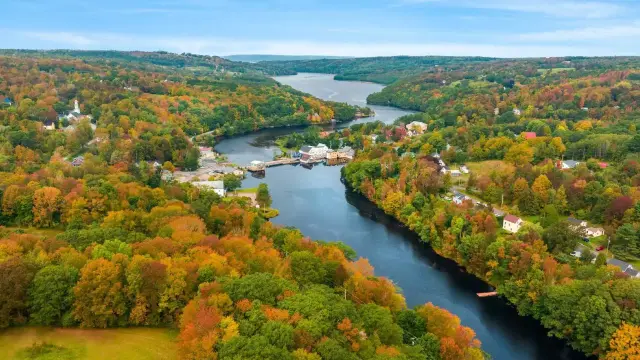 Aerial view of the Bear River in Annapolis County, Nova Scotia, during peak autumn foliage season. The river winds through rolling hills densely forested with colorful trees. A small village is visible on the river's edge.