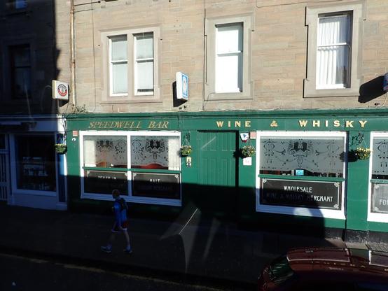 A green painted bar exterior on the ground floor of a stone tenement block on Perth Road Dundee. The pub windows are etched glass and advertise wines and whisky, as does the lettering above them.