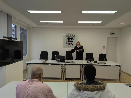 Gerichtssaaa.
Im Vordergrund sieht man die Rückenansicht von zwei männlich gelesenen Personen. Im Fokus in der Mitte ist eine Richterin in schwarzer Robe dabei ihren Stuhl hinter dem Richter:innen-Tisch weg zu schieben. Hinter ihr das Wappen von baden-Württemberg mit drei Löwen. Der Saal ist weiß und hell. Links steht ein Fernseher und mal sieht ein Fenster