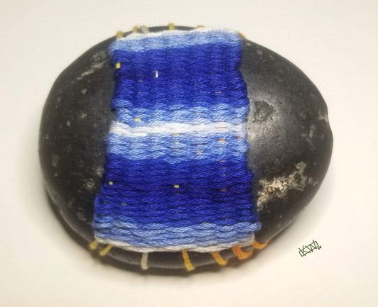 Photo of a black oval stone wrapped in variegated blue embroidery thread woven into a little blanket with stripes of dark blue, light blue, and white.