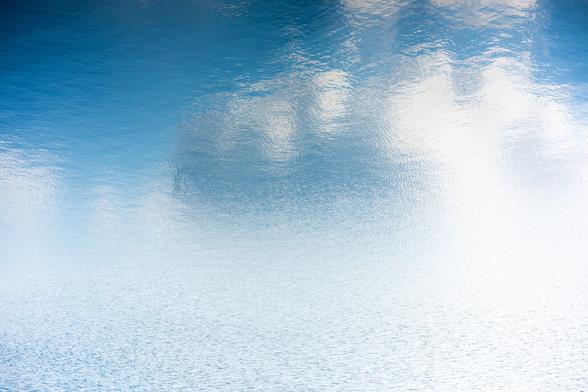 Rippling water surface reflecting a blue sky and white clouds, creating a textured, abstract pattern with gentle gradients from blue to white.