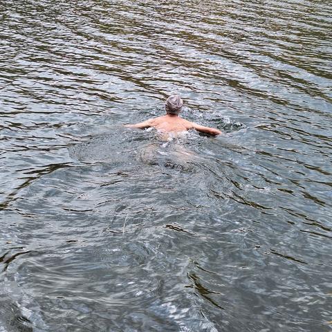 Der Kopf mit beige-braun gestreifter Mütze und Oberkörper eines Brust-Schwimmers von hinten. Die Arme sind seitlich ausgestreckt und bis zum Unterarm zu sehen. Die Wasseroberfläche ist vor dem Schwimmer in regelmäßigem Muster leicht gewellt und spiegelt im Wechselspiel von hell und dunkel die Farben des Himmels und der Umgebung von Blau bis Grau. Hinter dem Schwimmer ist das Wasser entsprechend verwirbelt in vorherrschend hellen Blautönen.