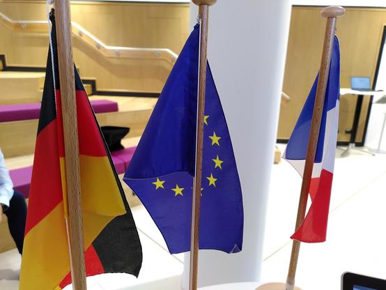 Auf einem Tisch in einem Veranstaltungsraum stehen eine deutsche, eine französische und eine europäische Fahne.