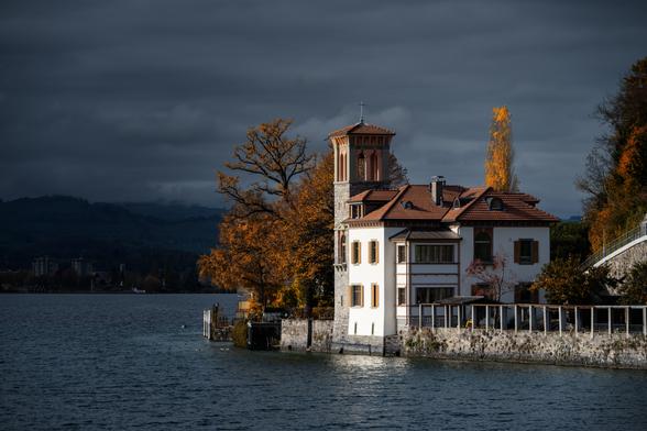 Ein markantes, historisches Gebäude am Ufer eines Sees unter einem dramatischen, dunklen Himmel. Das Gebäude ist im Stil eines toskanischen Landhauses gehalten, mit hellen, cremefarbenen Fassaden und einem Ziegeldach. Ein auffälliger, viereckiger Turm mit Rundbogenfenstern und einem kleinen Zeltdach erhebt sich an einer Ecke des Hauses. Braune Fensterläden und Zierleisten ergänzen die Farbgebung.

Das Gebäude steht direkt am Wasser, dessen Oberfläche dunkelblau bis graublau schimmert. Eine robuste Steinmauer mit Geländer säumt das Ufer neben dem Haus. Links neben dem Gebäude und dahinter sind grosse Laubbäume mit leuchtend orangen und gelben Herbstblättern zu sehen, die einen starken Kontrast zum dunklen Himmel und dem dunklen Wasser bilden. Auch rechts hinter dem Haus sind goldgelbe Blätter zu erkennen.

Im Hintergrund erstreckt sich die Küstenlinie mit weiteren Bäumen und Andeutungen von Bebauung bis zu den verschwommenen, dunklen Bergen am Horizont.