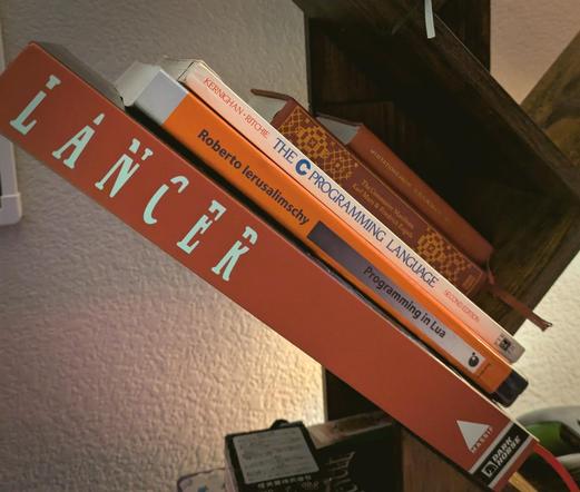 On a diagonal wooden bookshelf the following  are stacked: The Lancer TTRPG core book, Ierusalimschy's "Programming in Lua', Kernighan and Ritchie's 'The C Programming Language', Marx and Engels' 'The Communist Manifesto' and Quotations from Chairman Mao