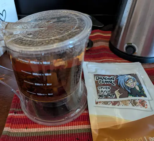 A photo of a tea infuser filled with steeping black tea, next to an empty pouch labeled "Dragon Eater", next to artwork of the character Momoyo Himemushi from Touhou Project.