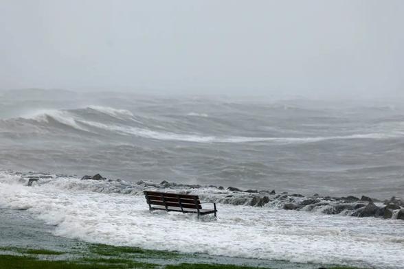 A wooden bench partially submerged in turbulent, frothy water along a shoreline, with dark storm clouds in the background.