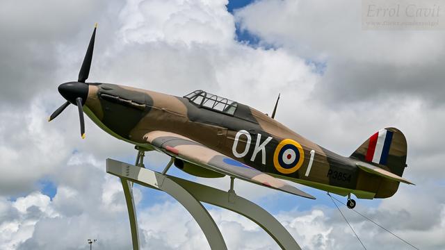 Full size replica World War 2 fighter mounted high on a stand seen from the side, banked towards us. It has green and brown camouflage, RAF markings, and the letters OK 1.