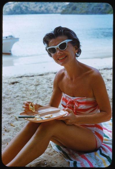 A smiling woman in a red and white striped dress sits on the beach holding a plate of food. She is wearing sunglasses, has painted nails, and her hair is styled into waves. In the background, there's a boat floating in calm blue waters with green hills visible along the horizon line. The photo appears to be faded or aged, suggesting it may have been taken many years ago.