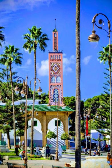 A vertical, vibrantly coloured photograph of a tall, ornate minaret tower featuring intricate geometric Zellige tiling in pink, blue, and yellow patterns. The minaret rises above a low, arched building with a green-tiled roof, set in a sunny public square framed by tall palm tree trunks and decorative street lamps. Several people are visible sitting on benches and walking in the square below, which features low green hedges and small trees, all under a bright blue sky.