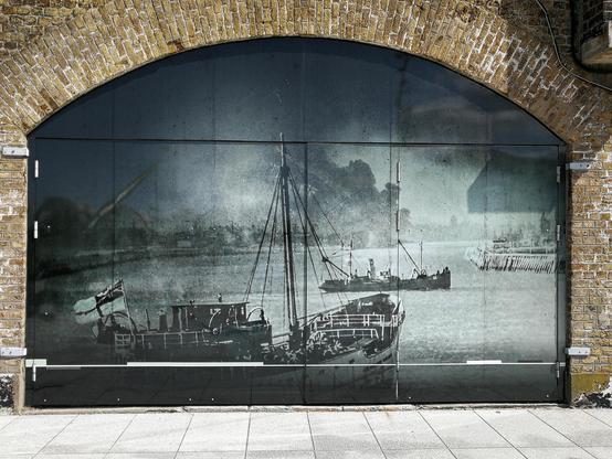 Large black-and-white historical image of boats displayed across a glass panel set within a brick archway. The photo features multiple ships, predominantly a large sailboat and a steamboat, depicted in an older maritime scene with industrial elements visible in the background. Distinctive urban elements like the light reflecting off the glass surface and the contrast with the aged brick surround are prominent.