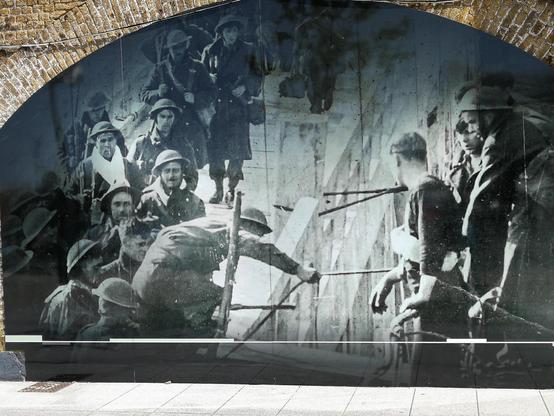 Black and white historical photo displayed on an arched outdoor mural showing a group of people in early 20th-century military attire, with some climbing a structure and others observing, creating a dynamic scene of activity and interaction.