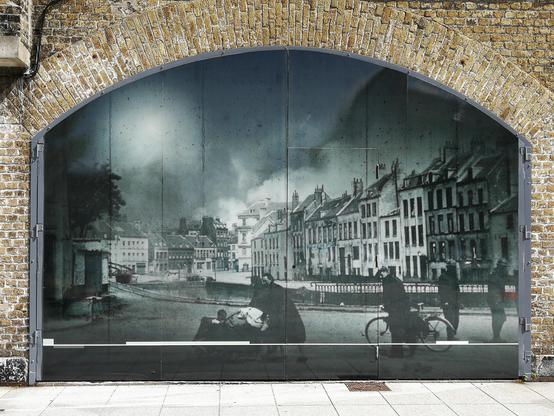 Large black and white photographic mural depicting a historic street scene, displayed on a semicircular wall under a brick archway. The mural features several people, some on bicycles and others walking, with row houses lining the street. Signs of weathering and age are visible on the surface of the mural.