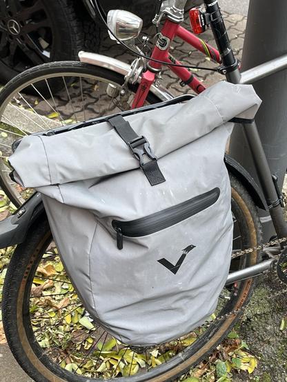 A grey roll-top bag with a small zip pocket is attached to a bicycle. The bag features a black logo, and the background includes parts of a pink bike and some fallen leaves.