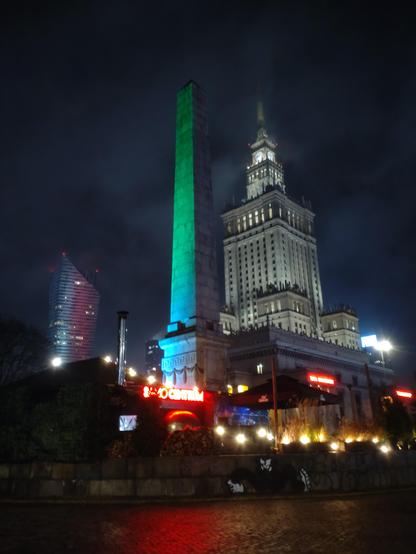 Same skyscraper from some distance, and in front of it stands Egyptian style obelisk washed in the green light