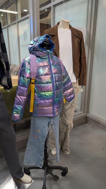 A child sized mannequin wearing a shiny, warm, water resistant purple and green coat and purple backpack and blue jeans; a second mannequin wearing tan pants, white shirt, and brown leather jacket