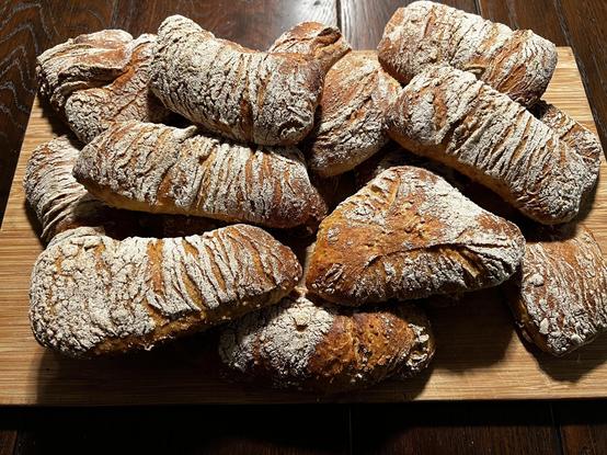 Auf einem dunklen Holztisch liegt ein Holzbrett. Darauf liegen zahlreiche frisch gebackene Brötchen