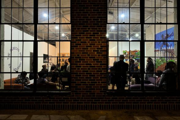 Photo taken from outside a brick building. Inside the two large windows is a large group of people discussing the importance of Assata Shakur as a revolutionary.