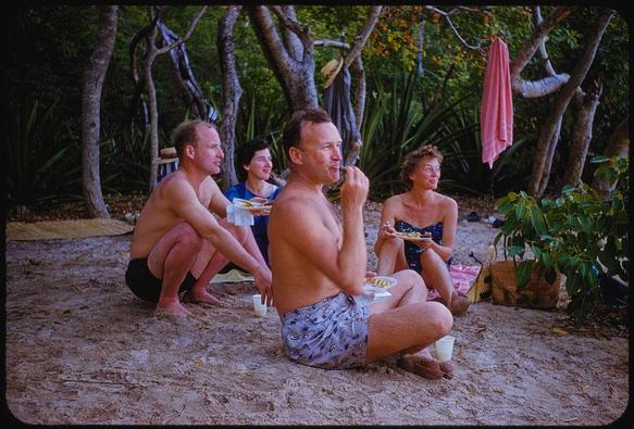 A group of people are gathered outdoors at a beachside picnic. The scene depicts four individuals, two men and two women, sitting on the sandy ground surrounded by trees with dense foliage in what appears to be a lush forested area. They are casually dressed for leisure time; one man is wearing patterned swim shorts while another has no shirt on but sports black swimming trunks. There's a variety of food items being enjoyed – some holding plates, cups and bowls that suggest they're partaking in a meal together.

Notably, the group displays relaxed postures typical of social gatherings during picnic moments: one man is caught mid-bite with his hand near his mouth while another gazes off into space. The women are similarly involved; their expressions range from engaged to contemplative as they interact or look around them.

Various objects contribute to this setting – a pink towel hangs on the tree, adding vibrant color contrast against the verdant backdrop. There's also evidence of picnic paraphernalia: a wicker basket containing items presumably related to food preparation and consumption sits beside one woman; cups are strewn about in close proximity suggesting casual dining.

The image carries an atmosphere that reflects both tranquility and conviviality, characteristic of Frissell’s portrayal within her "Way of Life" series which aimed at capturing candid life experiences. The photograph encapsulates a moment frozen i [...]