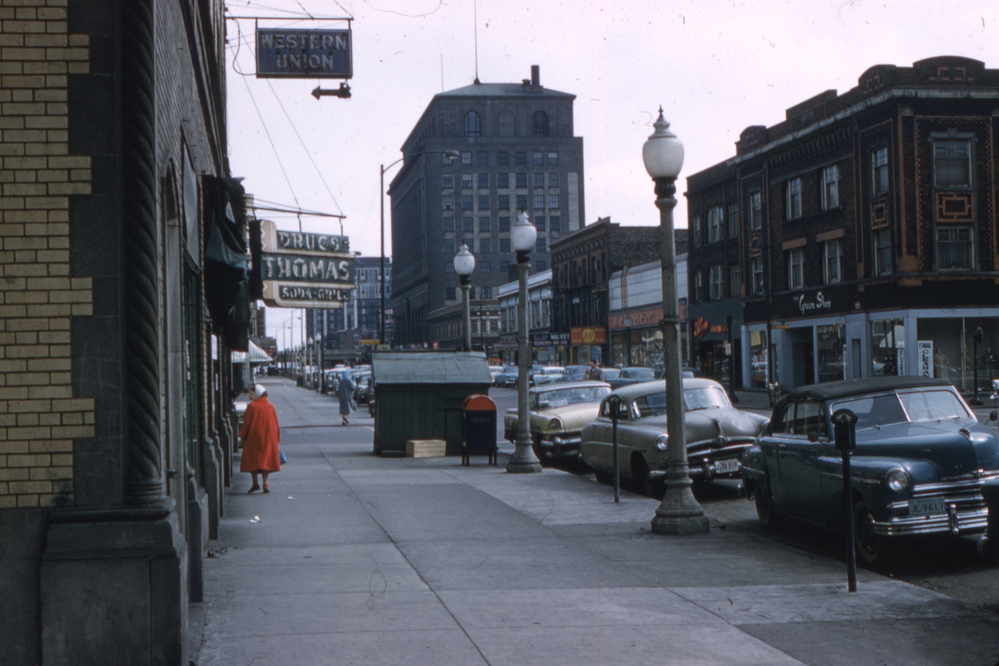 An old photograph of a city street somewhere in the States, featuring a woman in a red coat and old cars.
