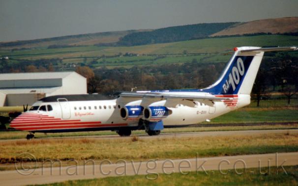 Side view of a high-winged, 4 engined jet airliner taxiing from right to left.
The plane is largely white, with a series of red stripes on the nose and forward fuselage, with more red stripes under the tail, and a dark blue belly, as well as a matt black patch on top of the forward fuselage, between the cockpit and the wing..
Then blue tail has diagonal , white "RJ100"titles .
The black registration "G-OIII" is on the lower rear fuselage.
The blue engine pods slung under the wing have red and white stripes at the bottom, and the text "LF507" on the outer pods.
Green grass lines a taxiway in the foreground, with more grass in the background leading up to a grey building in the background on the left, while trees give way to fields creeping up the side of a hill across the rest of the frame, under flat grey sky.