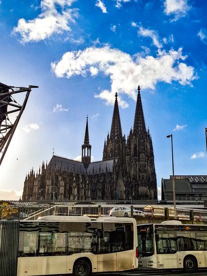 Der Kölner Dom dominiert die Szene unter einem sonnigen, wolkenverhangenen blauen Himmel. Im Vordergrund verdecken zwei weiße Stadtbusse einen Teil der erhöhten Straße. Die gotische Kathedrale ist majestätisch im Hintergrund positioniert.