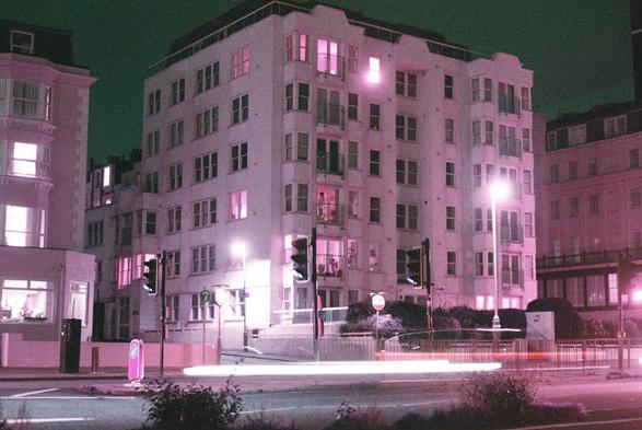 An apartment complex with light trails of cars on the street in front, with the whole scene hued purple/pink thanks to the film stock