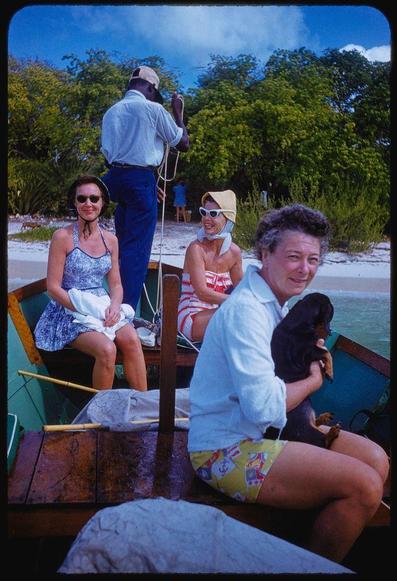 An image showing a group of people on board in the middle section of an ocean-going boat, with three women and one man visible. The photo seems to be taken during daytime hours under overcast skies as evidenced by the lack of harsh shadows.

The woman closest to us is seated wearing sunglasses, blue sleeveless top paired with dark skirt shorts while holding what appears to be a white towel or fabric in her lap; she has blond hair and wears large round glasses. The lady directly behind her seems to have black curly hair pulled back into a ponytail, donning red bikini bottom with the right half of body covered by an off-white sarong.

A third woman on our left is seated comfortably wearing sunglasses too, light skin tone with short dark brown hair, she's in blue sleeveless top and white skirt. A fourth lady next to her wears tan bucket hat, large round glasses reflecting bright sky as well; red and white striped bikini bottom covers upper body while holding what appears to be a towel or fabric around chest.

A man standing at the rear of boat with his back turned is wearing baseball cap, dark blue jeans paired with light blue shirt. He seems like crew member handling ropes on deck's stern area.