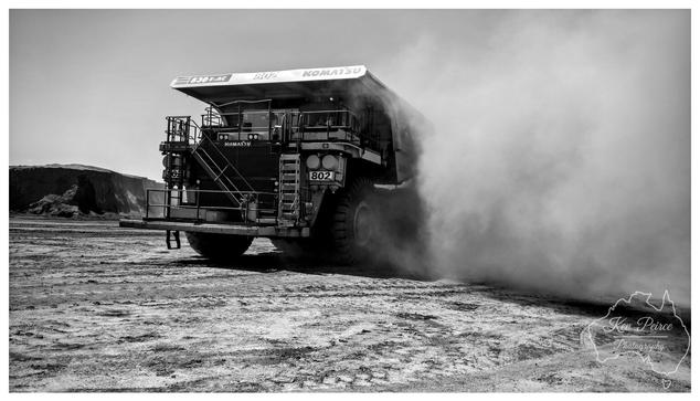 A dynamic, low angle, black and white photograph signed by Kev Peirce. A massive Komatsu 830E-AC haul truck, identified by its model number and '802' on the side, is seen from the rear quarter. The truck is driving on a dusty, cracked dirt surface, generating a huge, thick cloud of dust that billows dramatically to the right side of the frame. In the background, there is a large, sheer embankment of excavated material. The image captures the scale and activity of a major mining operation.